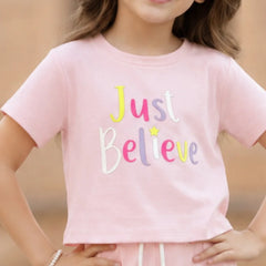 A young girl wearing a light pink short-sleeve crop top with the words 'Just Believe' in colorful embroidered letters, paired with matching pink pants.