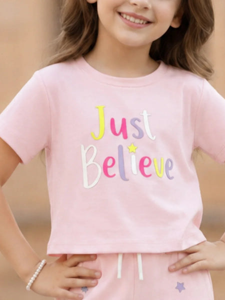 A young girl wearing a light pink short-sleeve crop top with the words 'Just Believe' in colorful embroidered letters, paired with matching pink pants.