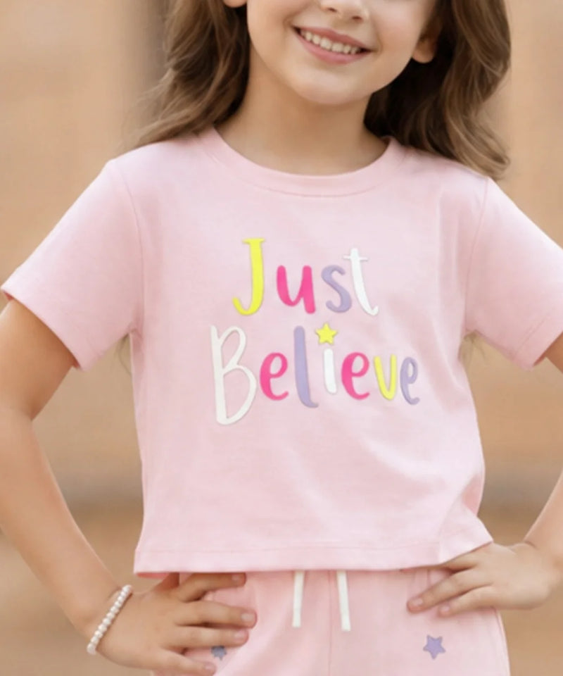 A young girl wearing a light pink short-sleeve crop top with the words 'Just Believe' in colorful embroidered letters, paired with matching pink pants.