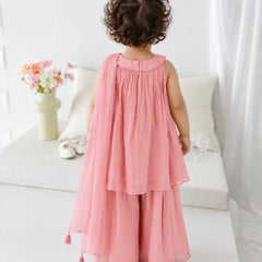 A toddler girl viewed from behind wearing a sleeveless dusty pink two-piece outfit with a pleated top and wide-leg skirt, standing in a bright, white-themed room.