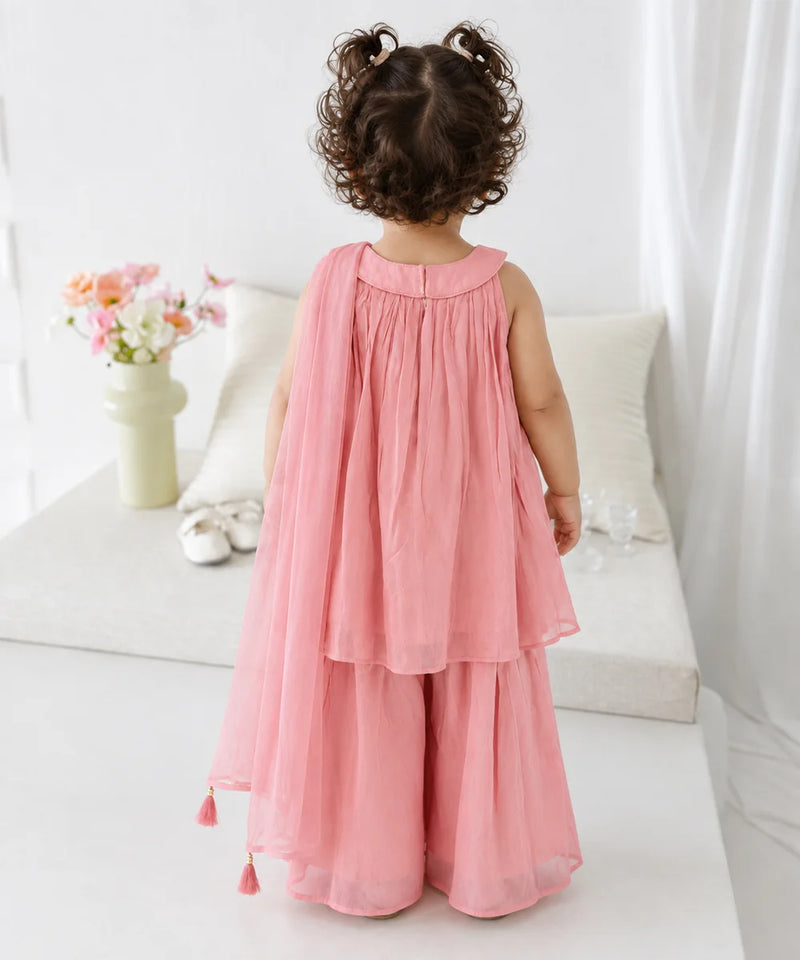 A toddler girl viewed from behind wearing a sleeveless dusty pink two-piece outfit with a pleated top and wide-leg skirt, standing in a bright, white-themed room.