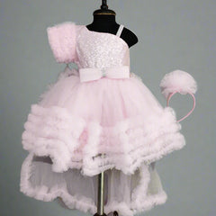 A pink knee-length frock with frill and bow detailing, displayed on a mannequin, including a detachable cape and a matching hair band.