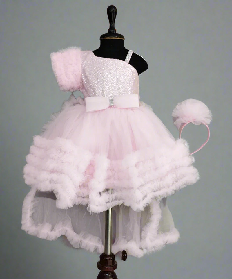 A pink knee-length frock with frill and bow detailing, displayed on a mannequin, including a detachable cape and a matching hair band.