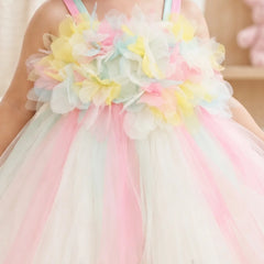 A close-up of a child wearing a pastel tulle dress with large layered flower appliqués across the chest in pink, yellow, mint, and white, plus thin pastel shoulder straps.