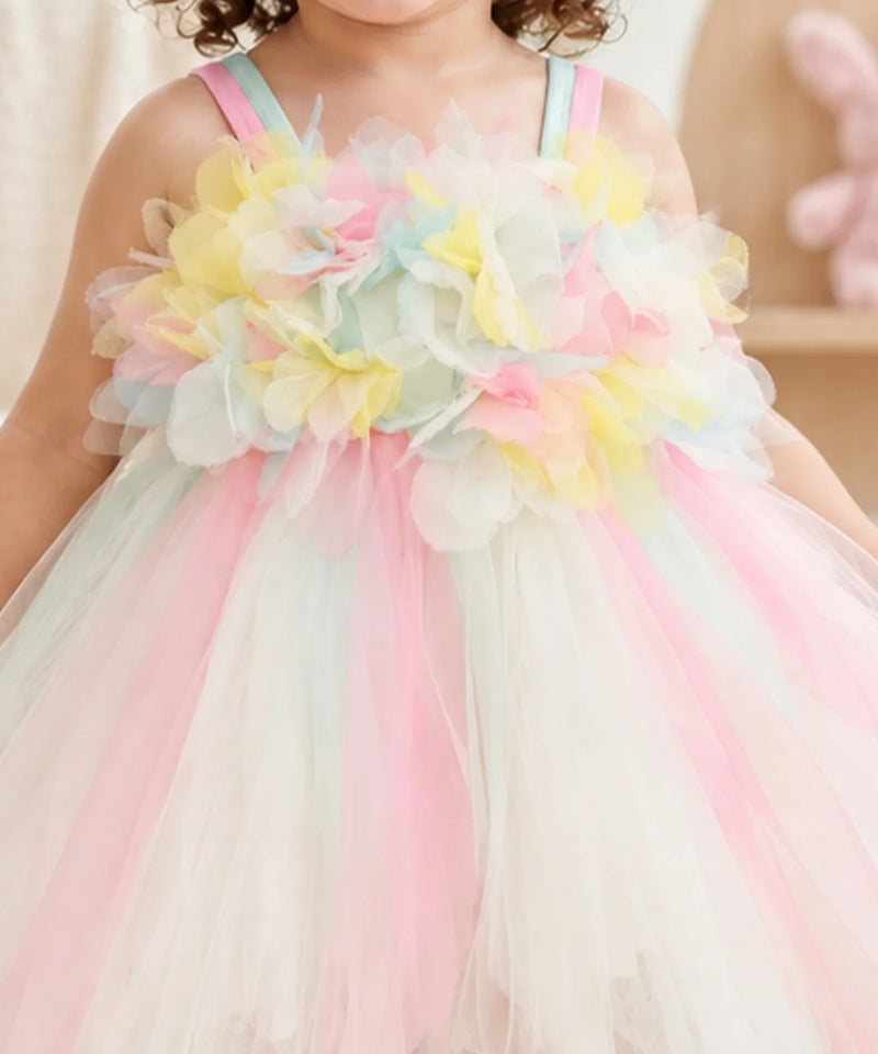 A close-up of a child wearing a pastel tulle dress with large layered flower appliqués across the chest in pink, yellow, mint, and white, plus thin pastel shoulder straps.