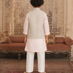 A child standing with their back to the camera, wearing a traditional Indian outfit consisting of a light beige waistcoat, a pale pink kurta, and white trousers. The setting is an ornate room with a patterned gold screen, a brown velvet sofa, and a patterned rug.