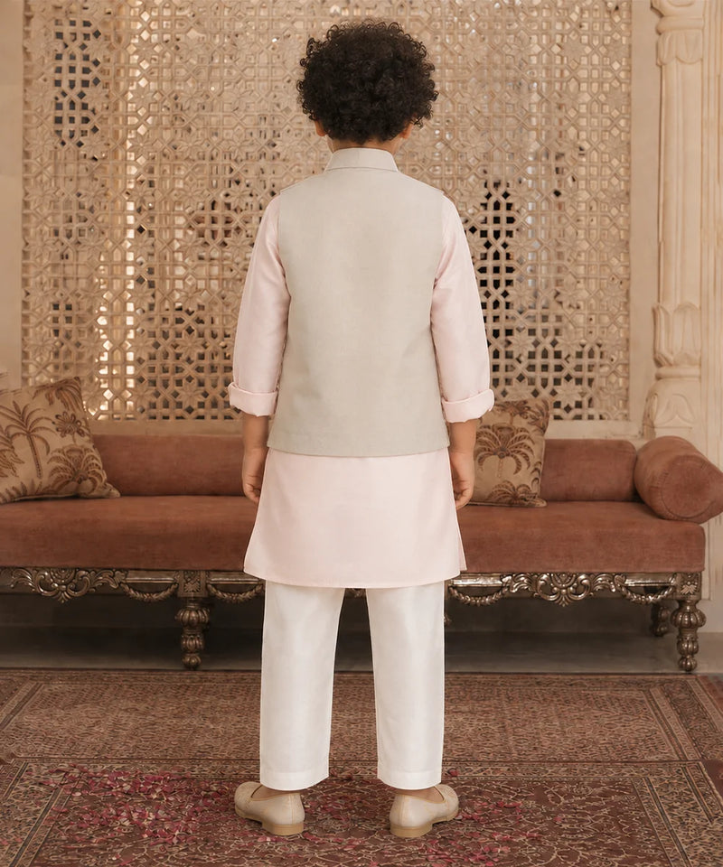 A child standing with their back to the camera, wearing a traditional Indian outfit consisting of a light beige waistcoat, a pale pink kurta, and white trousers. The setting is an ornate room with a patterned gold screen, a brown velvet sofa, and a patterned rug.