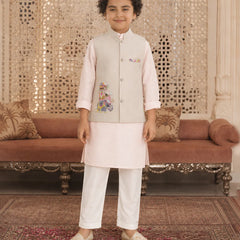 A young boy modeling a traditional Indian outfit consisting of a light beige embroidered vest, a pale pink kurta, white trousers, and beige shoes, standing in an ornate room with a patterned carpet and a brown sofa.