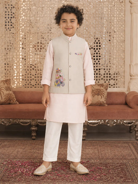 A young boy modeling a traditional Indian outfit consisting of a light beige embroidered vest, a pale pink kurta, white trousers, and beige shoes, standing in an ornate room with a patterned carpet and a brown sofa.