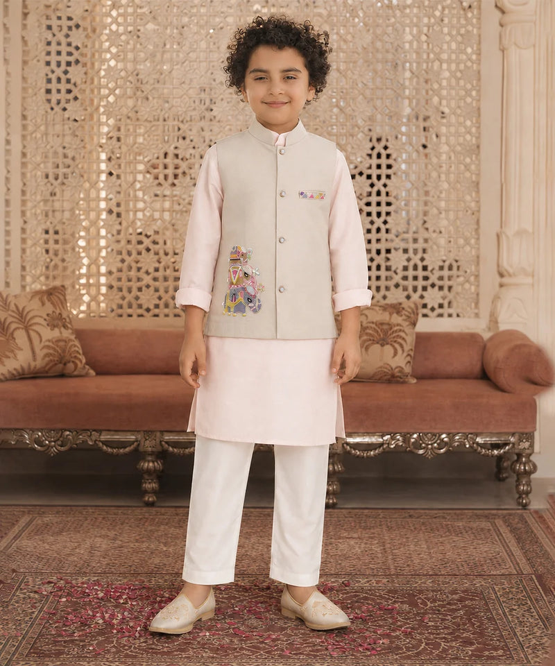 A young boy modeling a traditional Indian outfit consisting of a light beige embroidered vest, a pale pink kurta, white trousers, and beige shoes, standing in an ornate room with a patterned carpet and a brown sofa.