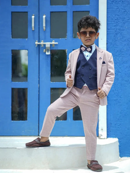 A boy wearing a pink check tuxedo with a white shirt, navy blue waistcoat, pocket square, and bow tie, standing in front of a blue door.