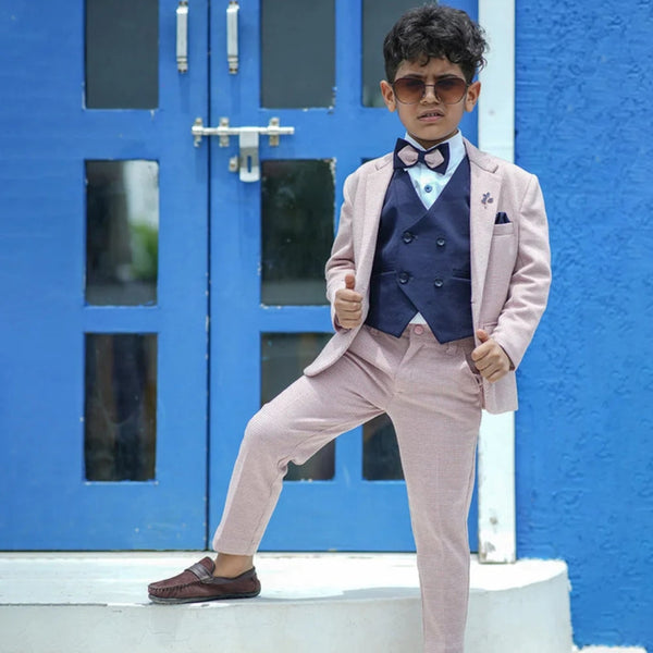 A boy wearing a pink check tuxedo with a white shirt, navy blue waistcoat, pocket square, and bow tie, standing in front of a blue door.