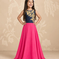 A young girl stands with hands on hips wearing a sleeveless formal dress with a navy blue bodice featuring gold floral embroidery and a flowing hot pink skirt. She is posing against a beige floral patterned backdrop.