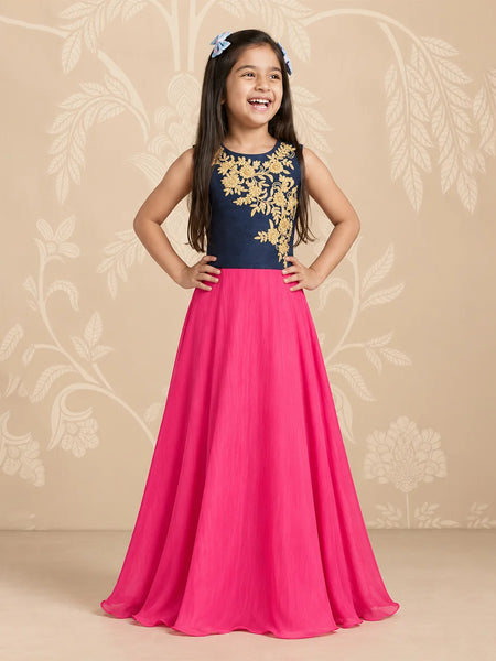 A young girl stands with hands on hips wearing a sleeveless formal dress with a navy blue bodice featuring gold floral embroidery and a flowing hot pink skirt. She is posing against a beige floral patterned backdrop.