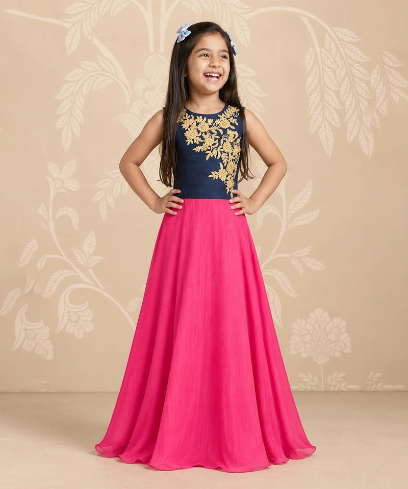 A young girl stands with hands on hips wearing a sleeveless formal dress with a navy blue bodice featuring gold floral embroidery and a flowing hot pink skirt. She is posing against a beige floral patterned backdrop.