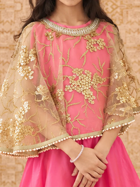 A child wearing a pink traditional outfit with a sheer gold embroidered cape-style top and a bright pink skirt, posed against a patterned beige background.