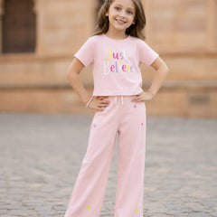 A young girl stands outdoors wearing a light pink two-piece outfit consisting of a short-sleeve t-shirt with 'Just Believe' printed on the front and matching wide-leg pants with small colorful embroidered details.