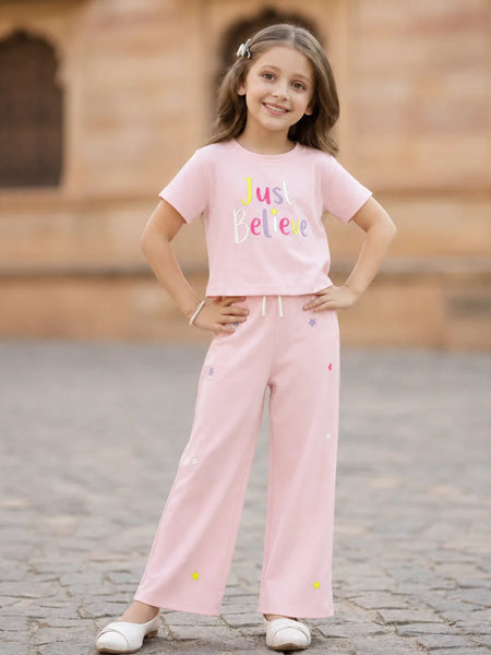 A young girl stands outdoors wearing a light pink two-piece outfit consisting of a short-sleeve t-shirt with 'Just Believe' printed on the front and matching wide-leg pants with small colorful embroidered details.