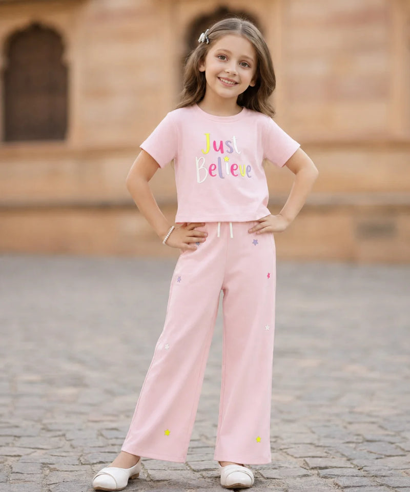 A young girl stands outdoors wearing a light pink two-piece outfit consisting of a short-sleeve t-shirt with 'Just Believe' printed on the front and matching wide-leg pants with small colorful embroidered details.