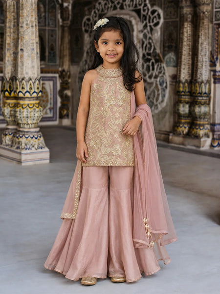 A young girl models a traditional Indian salwar kameez suit in dusty pink and gold. The set includes a sleeveless gold-embroidered kameez top, matching wide-leg pink trousers, and a sheer pink dupatta draped over her arm.