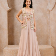 A young girl models a traditional Indian outfit consisting of a dusty pink wide-leg skirt, a matching pink crop top, and a sleeveless embellished vest in pink, gold, and green floral embroidery, set against a beige stone archway background.