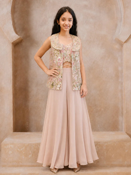 A young girl models a traditional Indian outfit consisting of a dusty pink wide-leg skirt, a matching pink crop top, and a sleeveless embellished vest in pink, gold, and green floral embroidery, set against a beige stone archway background.