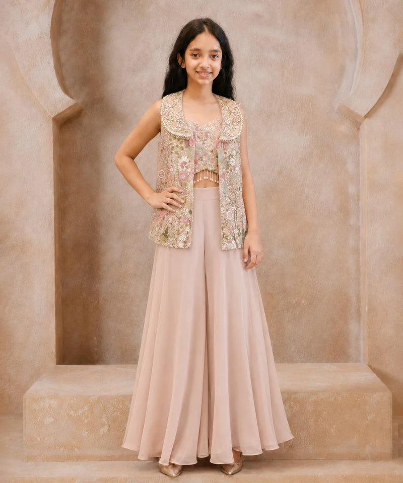 A young girl models a traditional Indian outfit consisting of a dusty pink wide-leg skirt, a matching pink crop top, and a sleeveless embellished vest in pink, gold, and green floral embroidery, set against a beige stone archway background.