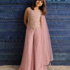 A young girl models a dusty pink traditional outfit consisting of a sleeveless embellished top, wide-leg pants, and a matching sheer dupatta, set against a textured blue patterned backdrop.