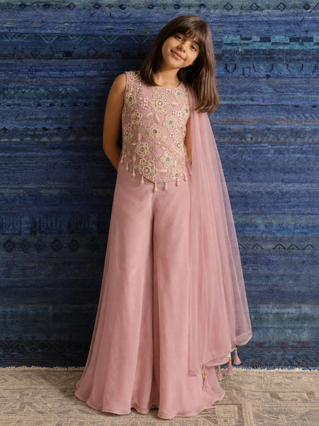 A young girl models a dusty pink traditional outfit consisting of a sleeveless embellished top, wide-leg pants, and a matching sheer dupatta, set against a textured blue patterned backdrop.