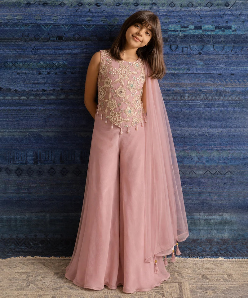 A young girl models a dusty pink traditional outfit consisting of a sleeveless embellished top, wide-leg pants, and a matching sheer dupatta, set against a textured blue patterned backdrop.