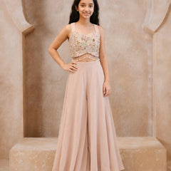 A young girl stands in a beige stone alcove wearing a two-piece formal outfit consisting of a heavily embroidered light pink and gold crop top with beaded fringe, and matching high-waisted blush pink wide-leg pants.