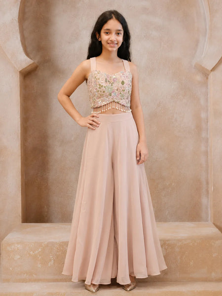 A young girl stands in a beige stone alcove wearing a two-piece formal outfit consisting of a heavily embroidered light pink and gold crop top with beaded fringe, and matching high-waisted blush pink wide-leg pants.