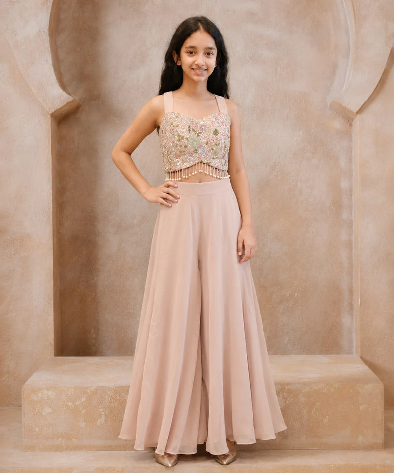 A young girl stands in a beige stone alcove wearing a two-piece formal outfit consisting of a heavily embroidered light pink and gold crop top with beaded fringe, and matching high-waisted blush pink wide-leg pants.