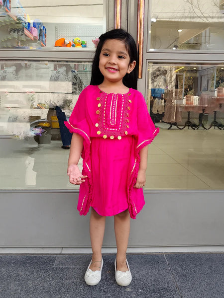 A girl wearing a pink kaftan style dress with embellishments, standing in front of a store display.