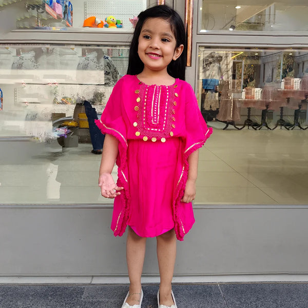 A girl wearing a pink kaftan style dress with embellishments, standing in front of a store display.