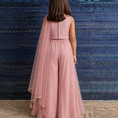A child model viewed from behind wearing a dusty pink sleeveless jumpsuit with a matching sheer tulle cape, standing against a textured blue and teal rug.