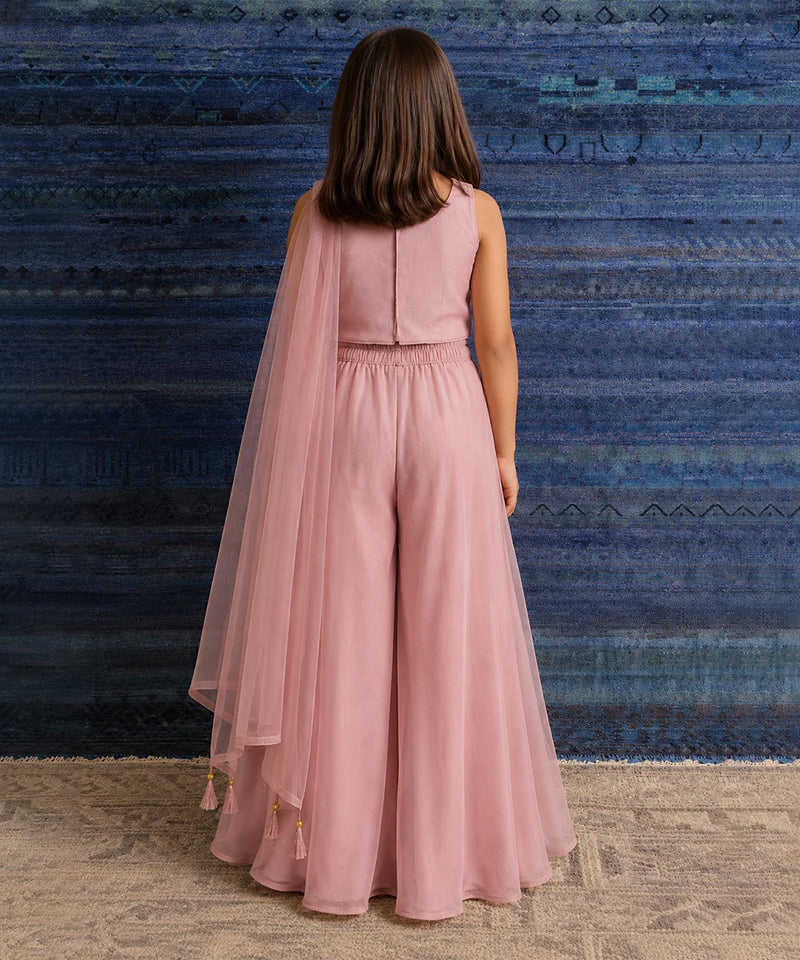 A child model viewed from behind wearing a dusty pink sleeveless jumpsuit with a matching sheer tulle cape, standing against a textured blue and teal rug.