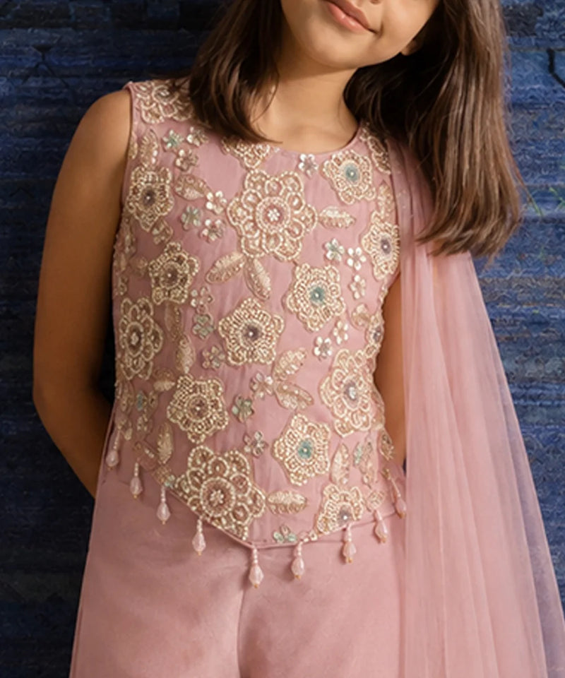 A child wearing a sleeveless dusty pink traditional outfit with intricate gold floral embroidery and beadwork, paired with a sheer pink dupatta, set against a dark blue textured background.