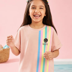 A young girl with long dark hair smiling and wearing a light pink t-shirt with vertical pastel stripes in blue, yellow, and green, and a smiley face graphic with the word 'HAPPY' on the front, set against a pink background.