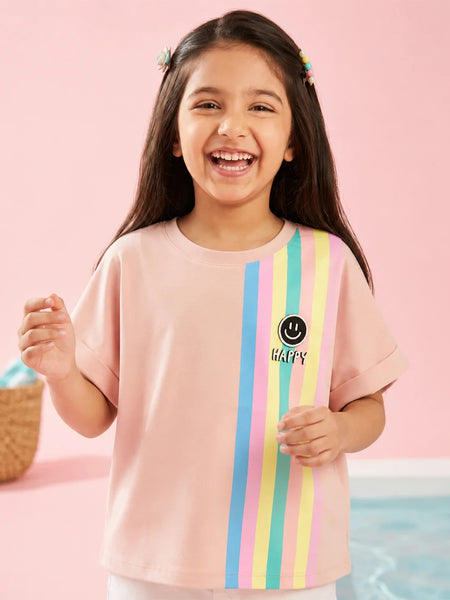 A young girl with long dark hair smiling and wearing a light pink t-shirt with vertical pastel stripes in blue, yellow, and green, and a smiley face graphic with the word 'HAPPY' on the front, set against a pink background.