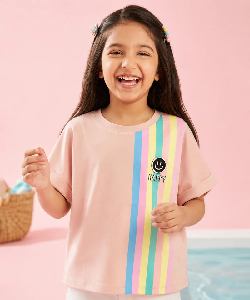 A young girl with long dark hair smiling and wearing a light pink t-shirt with vertical pastel stripes in blue, yellow, and green, and a smiley face graphic with the word 'HAPPY' on the front, set against a pink background.