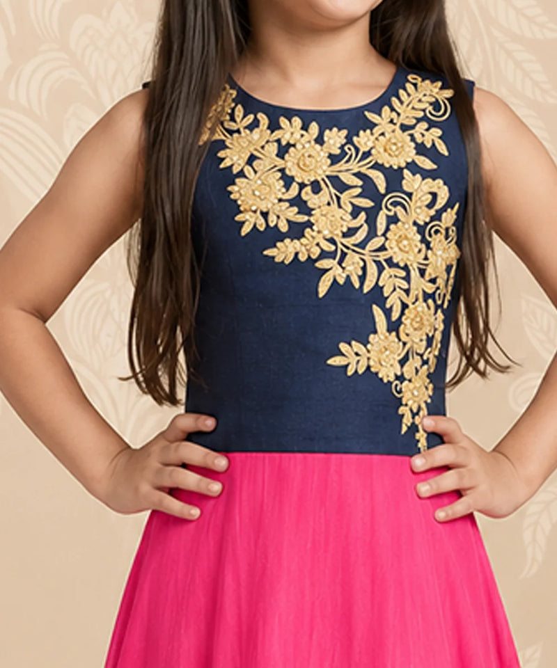 A sleeveless navy blue dress with intricate gold floral embroidery on the bodice, worn by a child with a bright pink skirt, set against a beige patterned backdrop.
