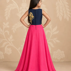 A young girl is shown from the back wearing a sleeveless navy blue and hot pink gown with gold embroidery on the back, standing against a beige floral patterned backdrop.