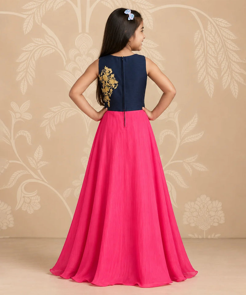 A young girl is shown from the back wearing a sleeveless navy blue and hot pink gown with gold embroidery on the back, standing against a beige floral patterned backdrop.