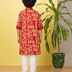 Person wearing a red and white patterned kurta with white pants, standing against a yellow wall with a plant.