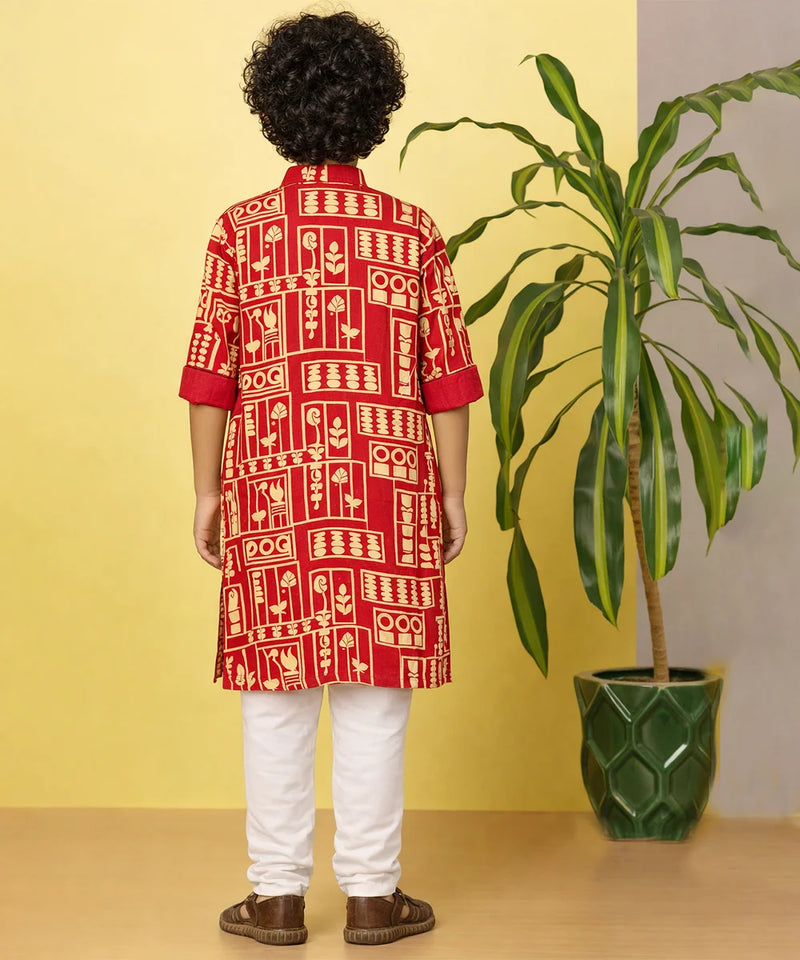 Person wearing a red and white patterned kurta with white pants, standing against a yellow wall with a plant.