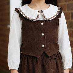 A child wearing a brown corduroy pinafore-style dress over a white long-sleeve blouse, shown from the neck to the hands against a brick wall background.