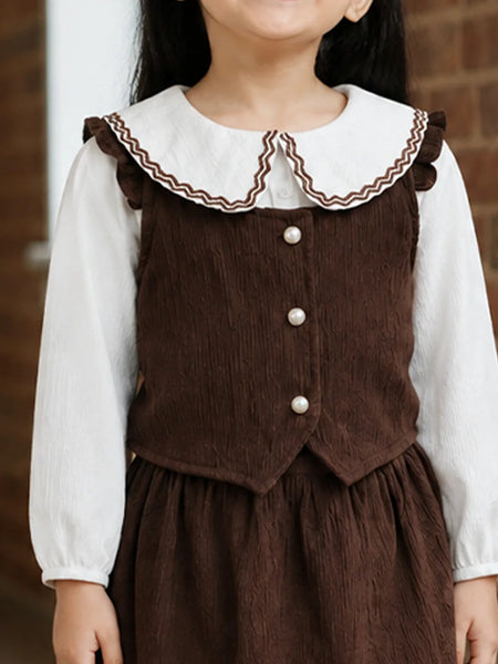 A child wearing a brown corduroy pinafore-style dress over a white long-sleeve blouse, shown from the neck to the hands against a brick wall background.
