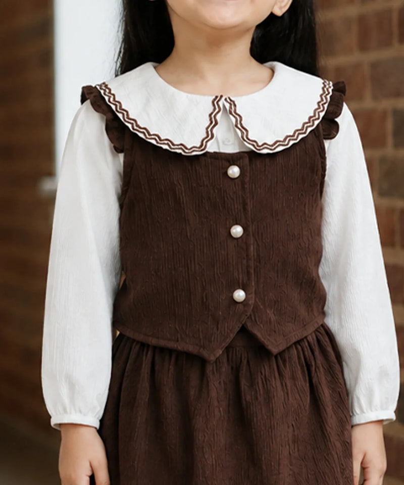 A child wearing a brown corduroy pinafore-style dress over a white long-sleeve blouse, shown from the neck to the hands against a brick wall background.