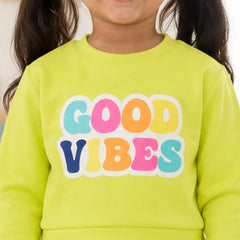 A close-up of a smiling child wearing a bright lime green sweatshirt with the words 'GOOD VIBES' printed across the chest in large, colorful bubble letters.
